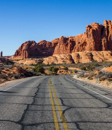 A beautiful shot of an empty road in the middle of a desert with bushes and cliffs in the distance under a blue sky